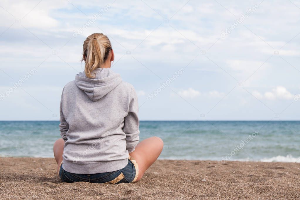 Warm clear sunny summer day. A girl in a gray sweatshirt and jeans shorts sits with her back to the camera on the sad shore and looks at the sea