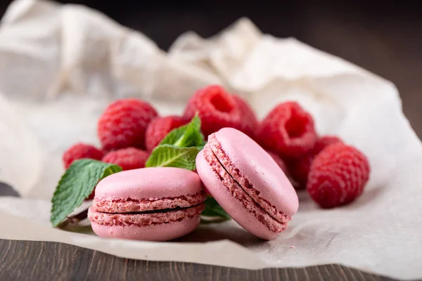 Pink macaroons with raspberries and mint leaves on a sheet of paper over a wooden background. — Stock Photo, Image