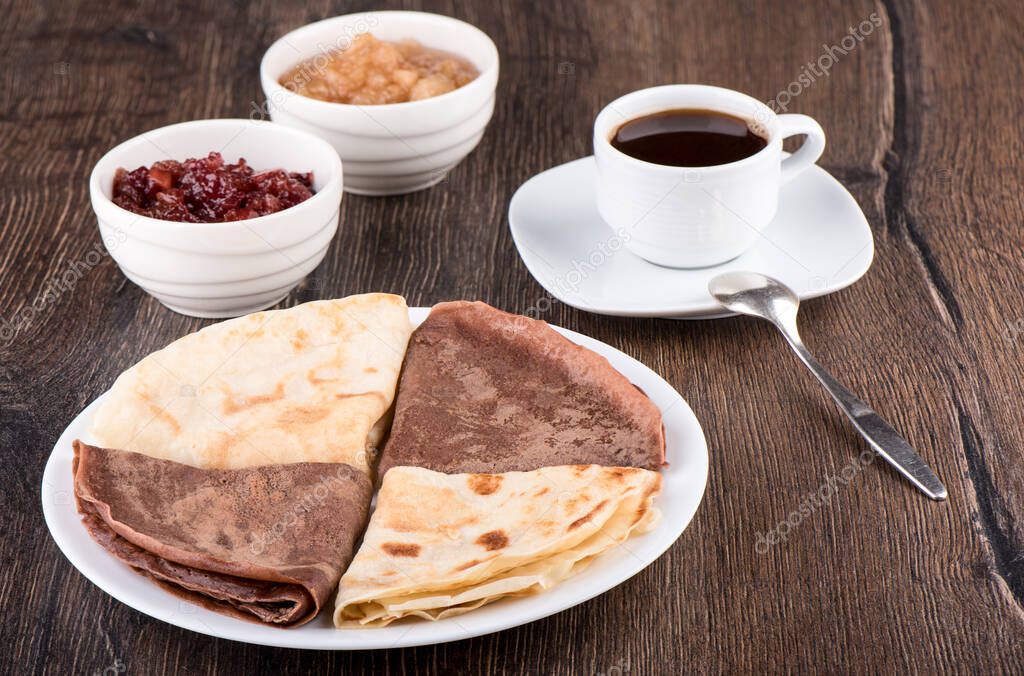 Pancakes are plain and with cocoa. Pancakes on a white plate, apple jam of different varieties and coffee over a wooden background. Traditional Russian breakfast.