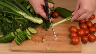 Kadın kesme tahtasının üzerinden yeşil soğanı makasla kesiyor. Tahta bir masada yeşiller, domatesler ve salatalıklar. Kırsal biçim.