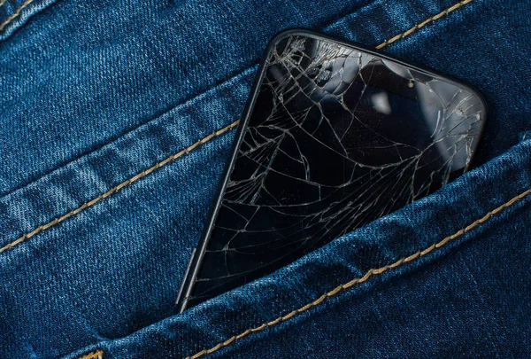 Mobile phone with a broken screen in the pocket of blue jeans.