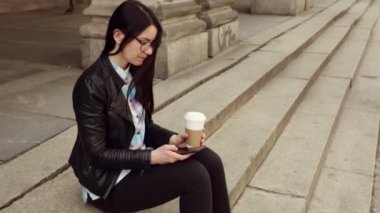 Güzel bir kız şehir adımda oturur ve onu smartphone ile görünüyor, kahve içer ve hayat sahiptir.