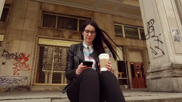 迷人的女孩在市中心 在黑色的皮革夹克和眼镜 浏览社会网络 在您的智能手机 — 图库视频影像