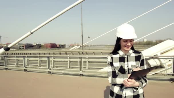 Žena v bílém stavbařskou přílbu a kostkovanou košili, stojící na visutý most a drží černou složku.