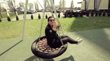 Neşeli güzel kız bir siyah deri ceket ve beyaz spor ayakkabı içinde bir salıncak üzerinde sürme. Güneşli bir günde salıncak.