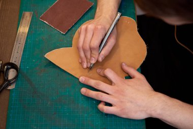 Ustanın erkek elleri gerçek deriden bir bıçak deseniyle kesiyor. Gerçek deri ve bıçakla çalışan deri tasarımcı ellerine yakın çekim. Tasarımcıların genel çalışma masası.