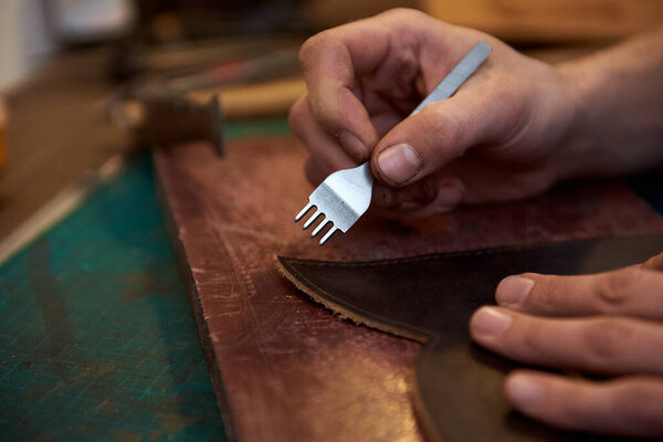 Человек, держащий в руках ремесленный инструмент и работающий с натуральной кожей за рабочим столом. Крупный план ремесленника, работающего с коричневой натуральной кожей с использованием ремесленных инструментов. Работа с натуральной кожей
.