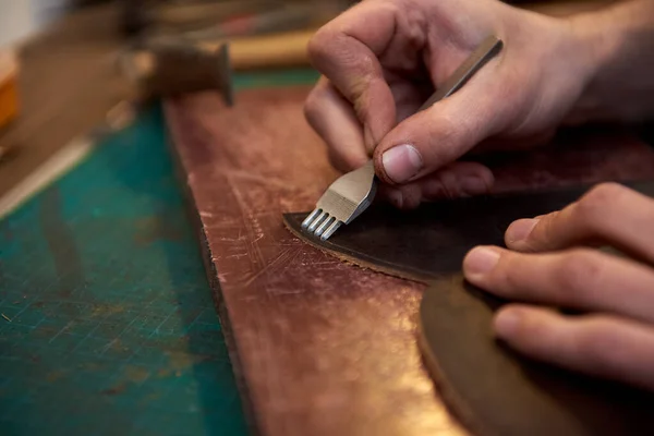 The leatherman holding crafting tool and working with brown genuine leather in his workshop. Closeup of craftsman working with natural leather using craft tools. Working with genuine leather.