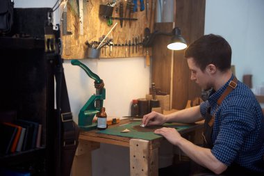 Deri ustası çalışma masasında doğal deri boyuyor. Gerçek deri boyama ustası. Mavi gömlekli deri bir zanaatkârın resmi masasında oturuyor ve hakiki kahverengi deriyle çalışıyor. Manufactory ve leat küçük işletme