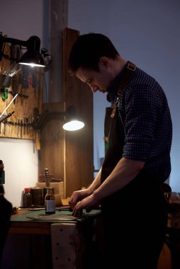 Masanın yanında duran ve hakiki kahverengi deriyle çalışan mavi gömlekli bir deri işçisinin dikey fotoğrafı. Fotoğraf, dikiş, el işi, deri ürünleri, hobiler, el yapımı ürünler, kendi küçük otobüsleri için kullanılabilir.