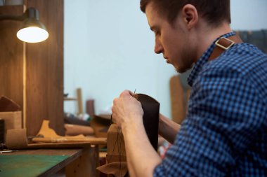 İğneyi iplikle tutan ve hakiki deriyle çalışan adam. Mavi gömlekli deri bir zanaatkârın resmi masasında oturuyor ve hakiki kahverengi deriyle çalışıyor. Bu fotoğraf, el işi ve dikiş işi için kullanılabilir.