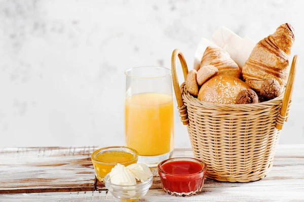 Hornear croissants desayuno y pan en cesta con jugo de naranja, miel, mantequilla, mermelada de frambuesa en la mesa de madera blanca — Foto de Stock