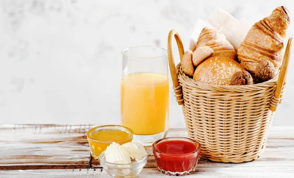 Hornear croissants desayuno y pan en cesta con jugo de naranja, miel, mantequilla, mermelada de frambuesa en la mesa de madera blanca —  Fotos de Stock