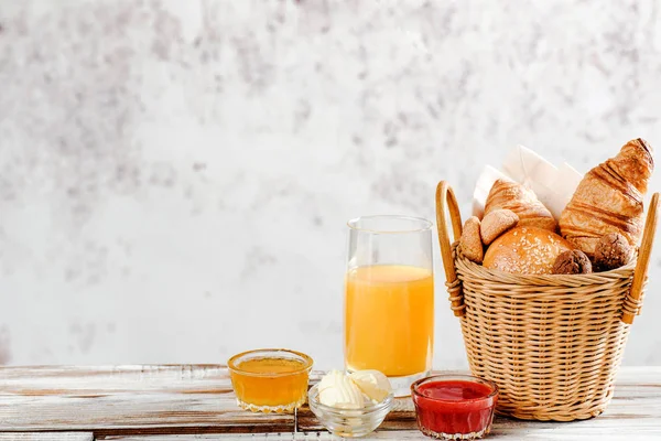 Hornear croissants desayuno y pan en cesta con jugo de naranja, miel, mantequilla, mermelada de frambuesa en la mesa de madera blanca —  Fotos de Stock