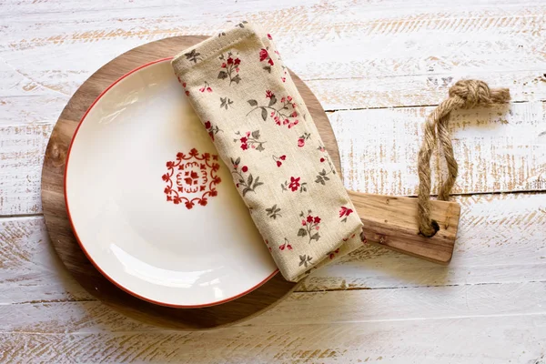 Off-white empty plate with red board on round cutting board, linen floral napkin, white plank wood background, Provence style, rural kitchen interior, minimalistic, top view