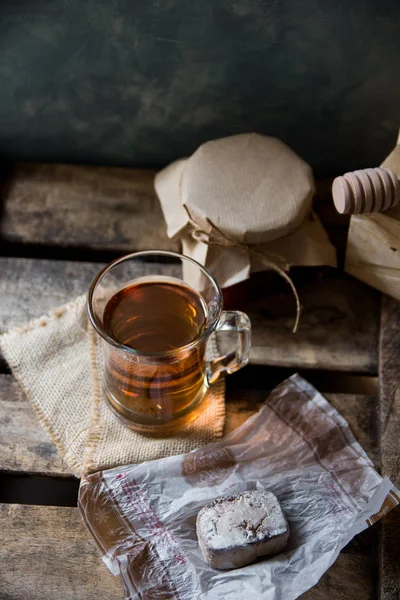 Glass cup with hot tea with jar of honey or jam, wood spoon, Spanish cookie polvoron on vintage box, grey wall background,rustic interior,cozy atmosphere — Stock Photo, Image