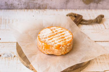 Whole wheel of soft French, German cheese with orange rind with mold on parchment paper, wood cutting board, concrete wall, rustic clipart