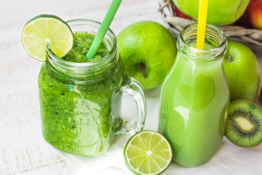 Kupa yeşil sebze güler yüzlü ve saman, elma, limon, mutfak masasında, güneş ışığı fleck kivi ile şişe meyve suyu ile jar