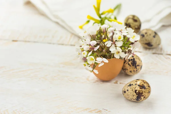 Pequeñas flores blancas amarillas de primavera en cáscara de huevo, huevos de codorniz, servilleta blanca sobre fondo de madera, decoración de Pascua, colores suaves — Foto de Stock