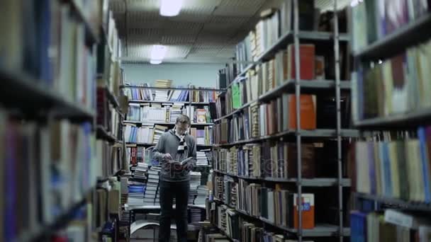 Estudiante leyendo un libro en la biblioteca, de pie entre filas, mover la cámara — Vídeo de stock