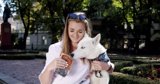 Ritratto bella giovane donna tenendo un cane husky tra le braccia e dando da mangiare al suo cane un panino. Colpo largo — Video Stock