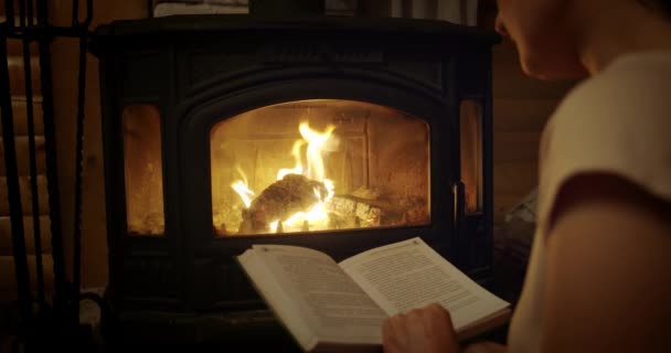 Movimiento lento, mujer leyendo un libro junto a la chimenea. Relajado concepto de vacaciones por la noche. Tiro medio. — Vídeo de stock