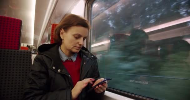 Wanita dewasa muda naik trem atau kereta api, melihat ke luar jendela dengan senyum samar. Dia memegang smartphone. Kelelahan yang menyenangkan setelah berjalan-jalan di kota, atau suasana hati yang bijaksana dan positif sebelum bekerja — Stok Video