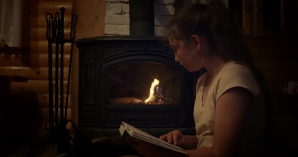 Acogedor, chica sentada junto a la chimenea, libro de lectura junto a la chimenea — Vídeo de stock