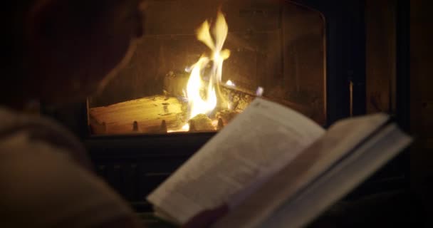 Útulné, dívka sedí u krbu, čte knihu u krbu, soustředí se na oheň — Stock video