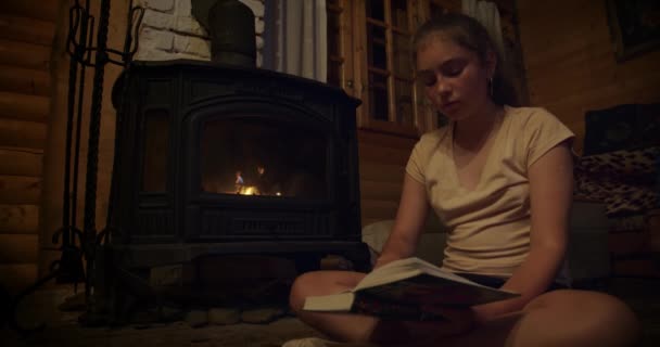 Acogedor, chica sentada junto a la chimenea, libro de lectura junto a la chimenea. de ancho tiro — Vídeos de Stock