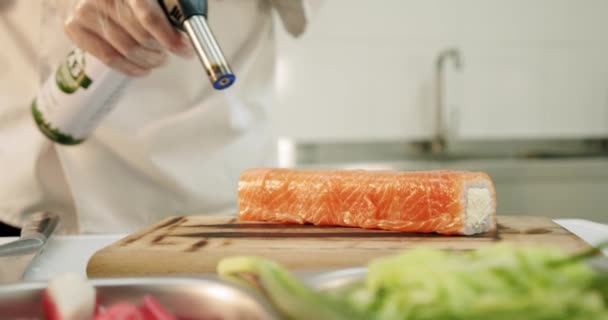 Cocina del restaurante. Chef de sushi masculino prepara rollos de sushi japonés de arroz, salmón, aguacate y nori. sushi se prepara con un quemador de antorcha de pistola de llama — Vídeo de stock