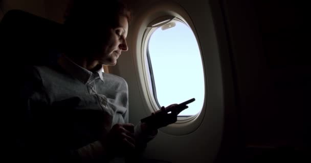 Increíble primer plano de la joven mujer relajada sosteniendo un teléfono inteligente en el vuelo del avión asiento de la ventana soleado. Movimiento lento — Vídeos de Stock