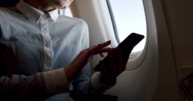 Akıllı telefonlu genç ve rahat bir kadının uçağın güneşli pencere kenarındaki bir pencereden fotoğrafını çekerken inanılmaz bir kare. Yavaş çekim yakın çekim