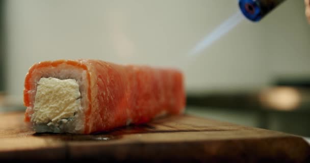 Lento movimentoRestaurante cozinha. O chef de sushi masculino prepara rolos de sushi japoneses de arroz, salmão, abacate e nori. sushi é preparado usando um queimador de chama arma tocha. Fechar . — Vídeo de Stock