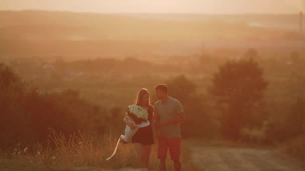 Pareja en el amor besos sosteniendo un ramo de flores sosteniendo un ramo de flores y caminando tomados de la mano en naranja puesta de sol entre las cabezas contra el paisaje montañoso — Vídeos de Stock