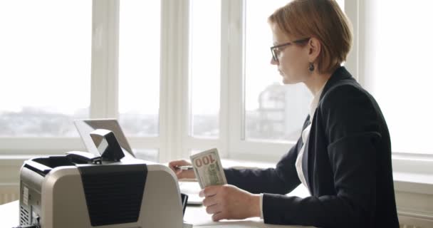 İş kadını, sayma makinesinde para sayıyor ahşap masada oturuyor ve sayma makinesinde bir tomar para sayıyor. — Stok video