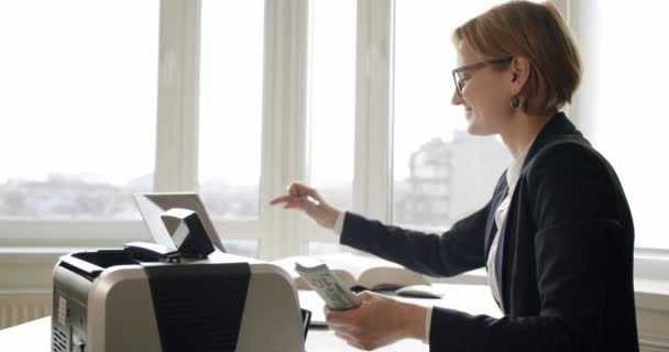 Heureuse femme d'affaires attrayante et souriante comptant l'argent comptant dans la machine de comptage assise au bureau en bois et comptant le grand faisceau de billets de dollar fosse dans la machine de comptage — Video