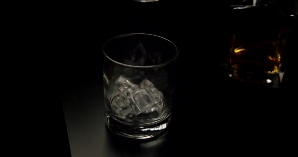 Повільний рух кидає лід у склянку пізніше золотистого віскі, що виливається в склянку з кубиками льоду з пляшки. крупним планом — стокове відео