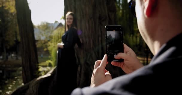 El hombre está haciendo un retrato fotográfico de una joven embarazada en un teléfono inteligente en el parque de la ciudad. De cerca. Movimiento lento — Vídeos de Stock