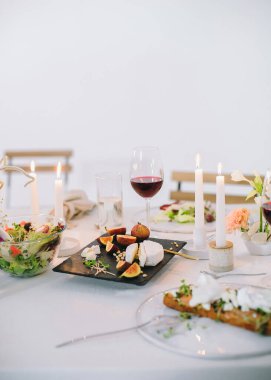 Tatil masasında çiçek buketi, şarap, mumlar, peynirli siyah kare tabak, çam fıstığı ve incir, salatalı cam tabak.