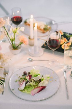 Çiçekli bir tatil masası, zeytinli ve çam fıstıklı salata, şarap, mum, kaşık, tabak, peçete