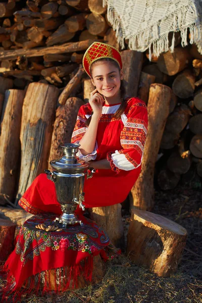 Vacker flicka i ryska folkdräkt sitter nära samovar och leende — Stockfoto