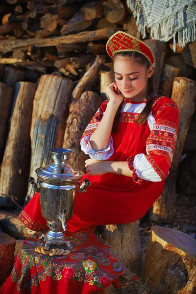 Krásná dívka v kroj sedí poblíž samovar a usmívá se — Stock fotografie