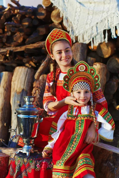 Две сестры в русских народных костюмах сидят рядом с самоваром и улыбаются — стоковое фото