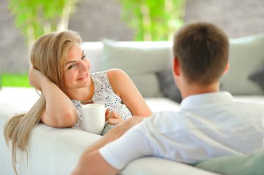 Kafasını bir el ve bir fincan lezzetli çay kocası seyir aşık başka bir elinde tutan bir kanepede oturan yakışıklı uzun saçlı sarışın genç kadın propped