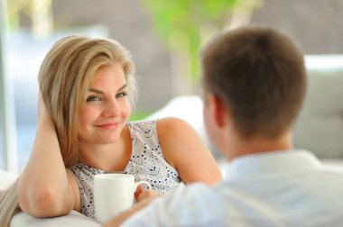 Kafasını bir el ve bir fincan lezzetli çay kocası seyir aşık başka bir elinde tutan bir kanepede oturan yakışıklı uzun saçlı sarışın genç kadın propped