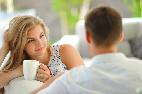 长发漂亮坐在沙发上的年轻金发女人撑起她的头，一手拿着一杯美味的茶，在另一只手，看着她的丈夫的爱 — 图库照片