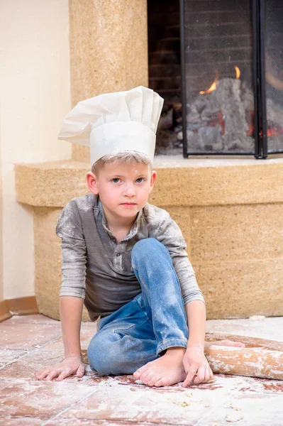 Un niño con sombreros de chef cerca de la chimenea sentado en la cocina f — Foto de Stock