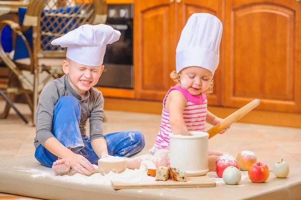 Dois irmãos - menino e menina - em chapéus de chef perto da lareira sentados no chão da cozinha sujos de farinha, brincando com comida, fazendo bagunça e se divertindo — Fotografia de Stock
