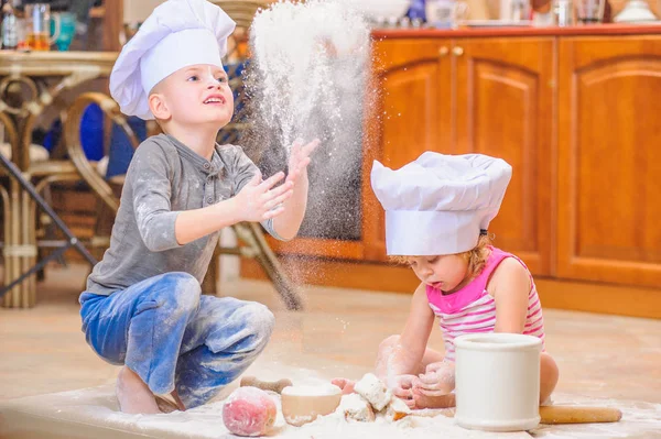 Two siblings - boy and girl - in chef's hats sitting on the kitc — Stock Photo, Image